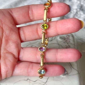 May include: A gold-toned bracelet featuring oval-shaped gemstones in shades of orange, green, purple, and blue. The bracelet is held between fingers, showcasing its design and color variations. The gemstones are set in a linear pattern.