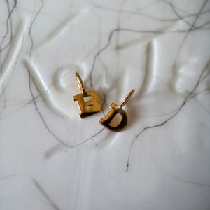 May include: Two small gold-toned metal letter charms, one shaped as the uppercase letter B and the other as the lowercase letter d. Each charm features a small attached jump ring at the top for hanging, with a polished, reflective metallic surface.