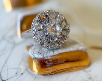 May include: A silver ring with a large, round cluster of clear gemstones. The ring is set on a small, white ceramic tile with gold trim. The tile has a black, web-like pattern.