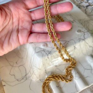 May include: A gold-colored rope chain necklace. The necklace is displayed on a white marble tray with gray veining. The chain is made of twisted metal links, creating a rope-like texture. The necklace is a classic jewelry piece.