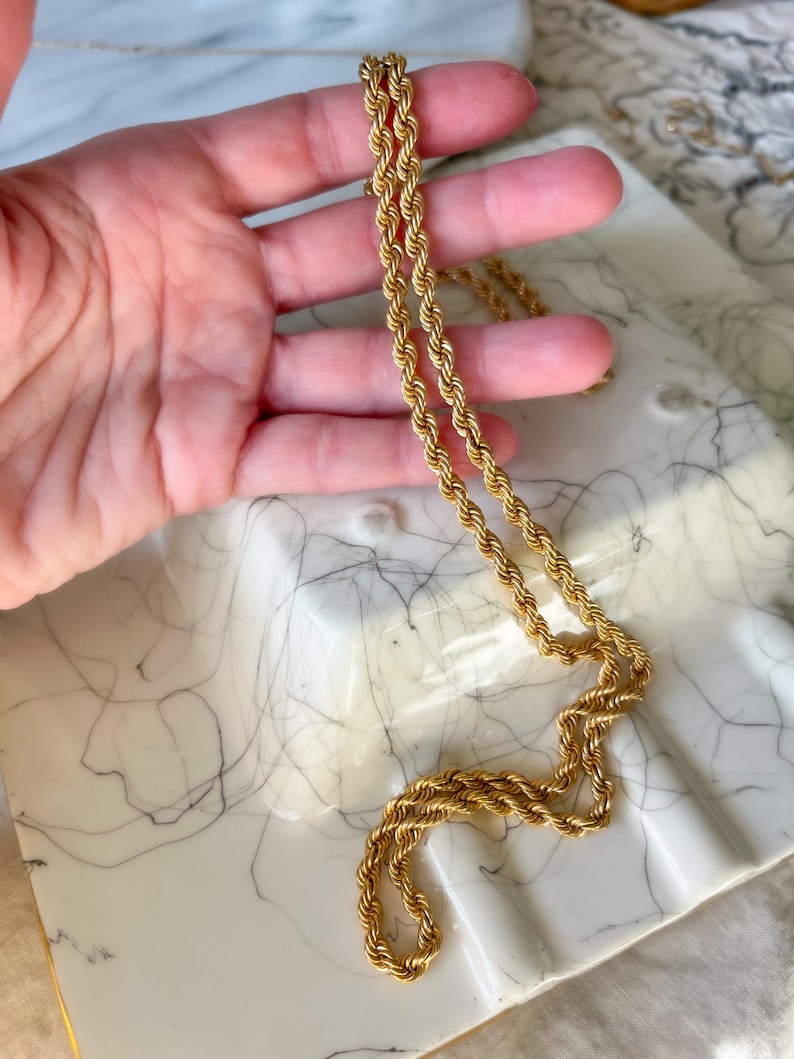 May include: A gold-colored rope chain necklace. The necklace is displayed on a white marble tray with gray veining. The chain is made of twisted metal links, creating a rope-like texture. The necklace is a classic jewelry piece.