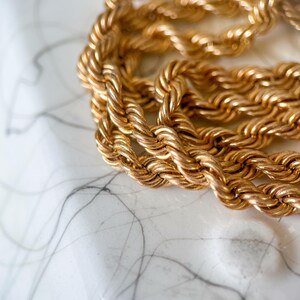 May include: Close-up of a gold-colored rope chain necklace. The chain is thick and appears to be made of twisted metal links. The background is a white surface with gray veining.