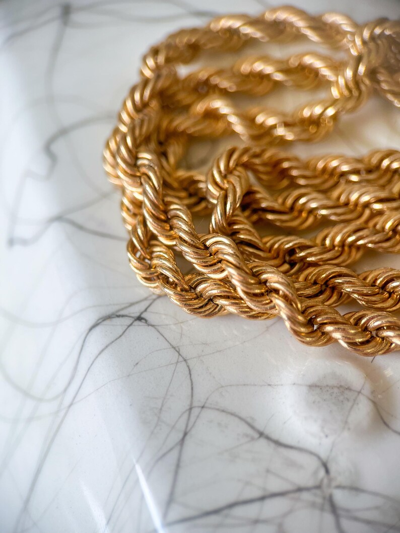 May include: Close-up of a gold-colored rope chain necklace. The chain is thick and appears to be made of twisted metal links. The background is a white surface with gray veining.