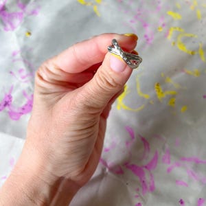May include: A silver-colored ring with a crisscross design, adorned with small, clear gemstones. The ring is held up against a background with purple and yellow paint splatters. The ring is a piece of jewelry.