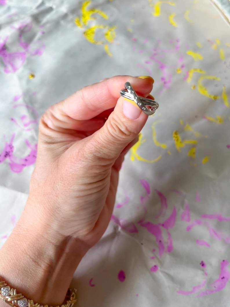 May include: A silver-colored ring with a crisscross design, adorned with small, clear gemstones. The ring is held up against a background with purple and yellow paint splatters. The ring is a piece of jewelry.
