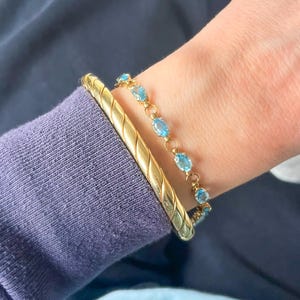 May include: A gold bracelet with a textured design and a delicate gold bracelet with oval-shaped blue gemstones. The bracelets are worn together on a wrist, against a backdrop of a blue garment.