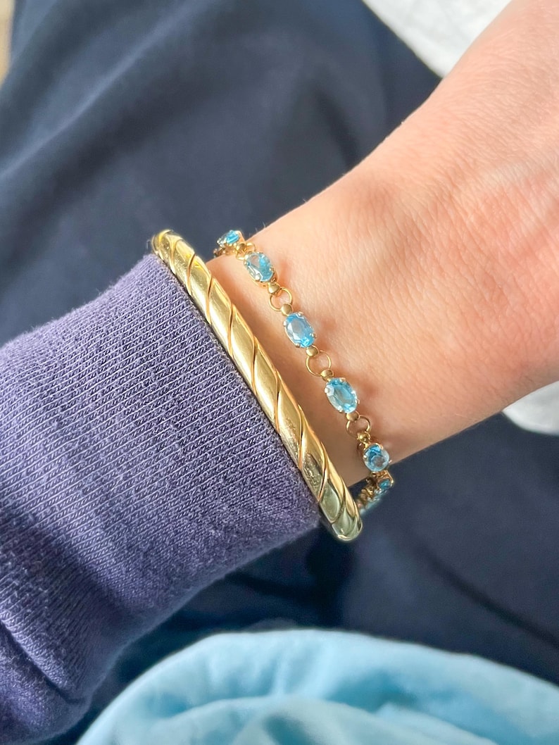 May include: A gold bracelet with a textured design and a delicate gold bracelet with oval-shaped blue gemstones. The bracelets are worn together on a wrist, against a backdrop of a blue garment.