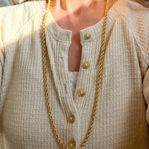 May include: A long, gold-colored rope chain necklace. The necklace is draped over a cream-colored waffle-knit cardigan with round, light brown buttons. The cardigan is worn over a white top.