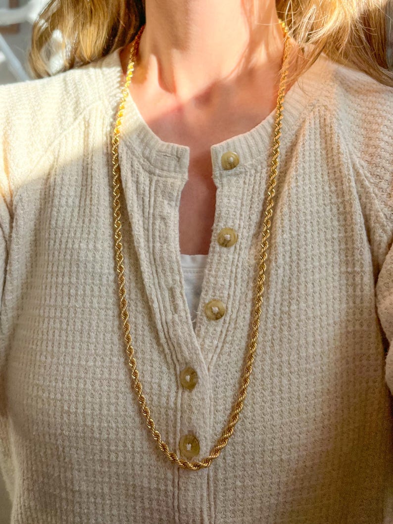 May include: A long, gold-colored rope chain necklace. The necklace is draped over a cream-colored waffle-knit cardigan with round, light brown buttons. The cardigan is worn over a white top.