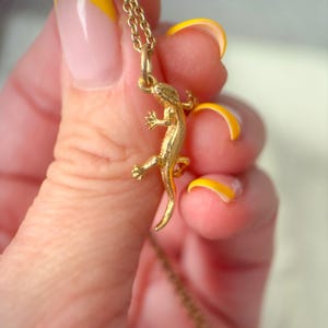 May include: A gold-colored lizard pendant necklace. The lizard charm is detailed with tiny legs and a curled tail, suspended from a delicate chain. The necklace is held up by a person with yellow-tipped nails.