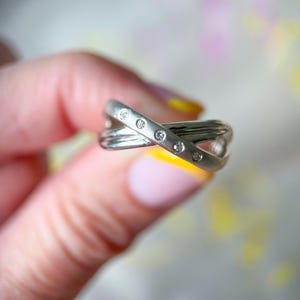 May include: A silver-toned ring with a crisscross design, featuring small, clear gemstones. The ring has a polished finish and is held against a soft, blurred background. The gemstones are evenly spaced along the top band of the ring.