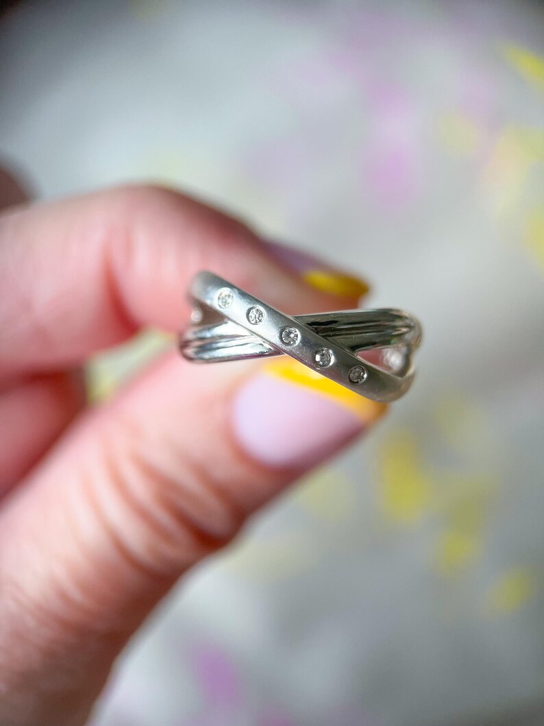 May include: A silver-toned ring with a crisscross design, featuring small, clear gemstones. The ring has a polished finish and is held against a soft, blurred background. The gemstones are evenly spaced along the top band of the ring.