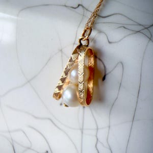 May include: A gold pendant necklace featuring two white spherical pearls encased within a textured, open-frame gold cage design. The pendant hangs from a delicate gold chain, showcasing a detailed, etched pattern on the metal frame surrounding the pearls.