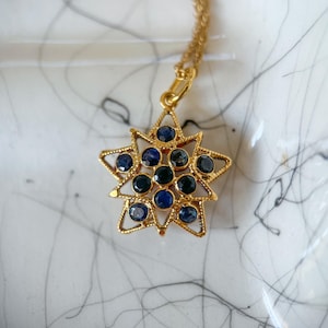 May include: A gold-colored star pendant necklace with a delicate chain. The star pendant is adorned with multiple dark blue gemstones. The necklace is displayed on a white surface with black markings.