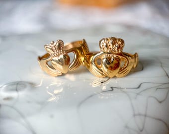 May include: Two gold-colored Claddagh rings. Each ring features a heart held by two hands, topped with a crown. The rings are displayed on a white and gray marbled surface, with soft lighting.
