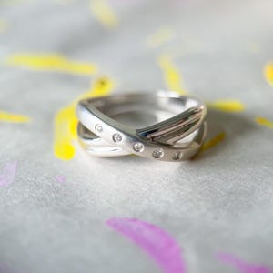 May include: A silver ring with a crisscross design, set with small, clear gemstones. The ring is displayed on a surface with colorful paint splotches of yellow and purple.