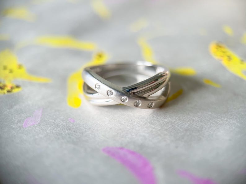 May include: A silver ring with a crisscross design, set with small, clear gemstones. The ring is displayed on a surface with colorful paint splotches of yellow and purple.