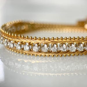 May include: Close-up of a gold bracelet with a repeating pattern of small, clear gemstones. The bracelet has a woven design with a series of small, round gold accents. The gemstones are set in a row along the center of the bracelet.