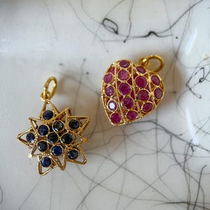 May include: Two gold-toned pendants. One is a starburst shape with dark blue and black stones. The other is a heart shape, filled with red stones. Both have small loops for hanging.