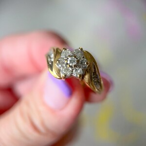May include: A gold ring featuring a cluster of small diamonds in a flower shape. The ring has a gold band with additional small diamonds set on the sides. The ring is held up against a soft, blurred background.