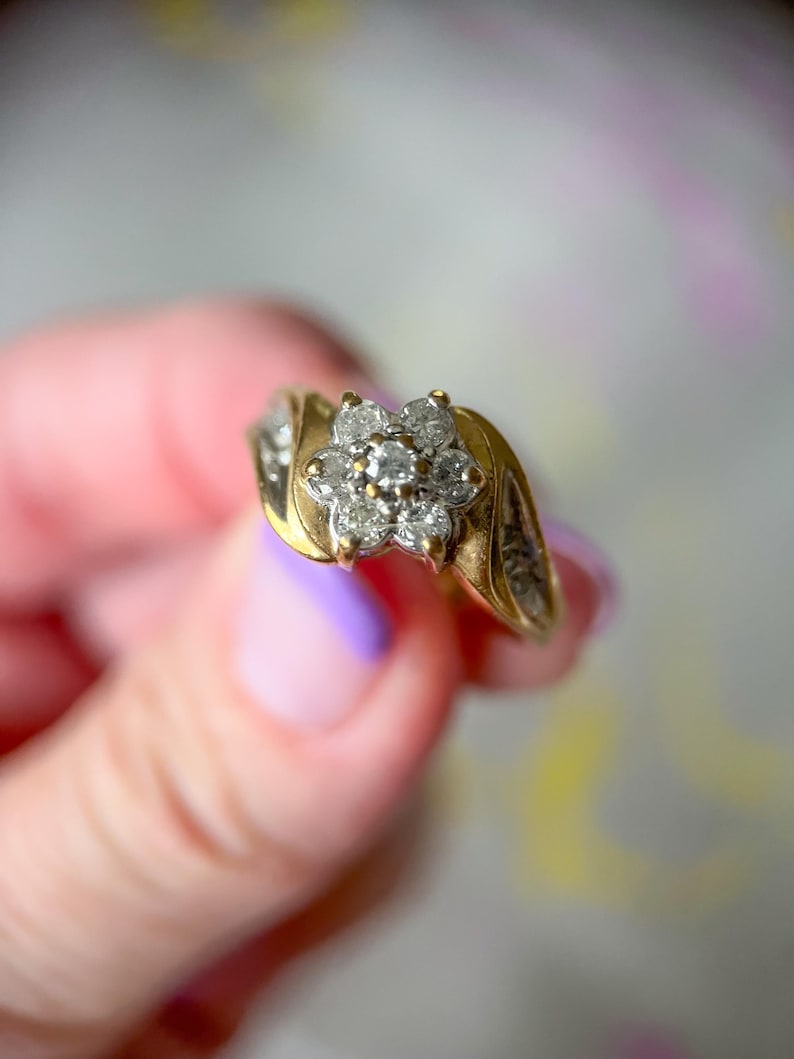 May include: A gold ring featuring a cluster of small diamonds in a flower shape. The ring has a gold band with additional small diamonds set on the sides. The ring is held up against a soft, blurred background.