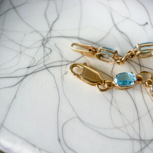 May include: Close-up of a gold bracelet with oval, light blue gemstones. The bracelet is on a white surface with black and gray abstract lines. The clasp is visible.