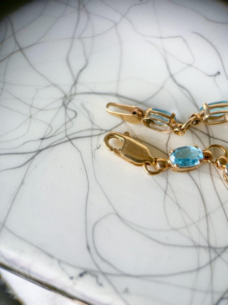 May include: Close-up of a gold bracelet with oval, light blue gemstones. The bracelet is on a white surface with black and gray abstract lines. The clasp is visible.