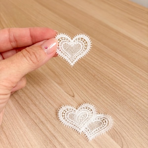 May include: Two white heart-shaped lace appliqués featuring a delicate, intricate woven grid pattern in the center and a scalloped, picot-style edge. The lace hearts are crafted with fine thread, showcasing a detailed, openwork design.