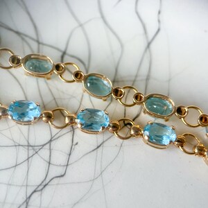 May include: Close-up of two gold-colored bracelets with oval, light blue gemstones. The gemstones are set in a gold-colored metal with small, circular accents. The bracelets are displayed on a white surface with a gray, abstract pattern.
