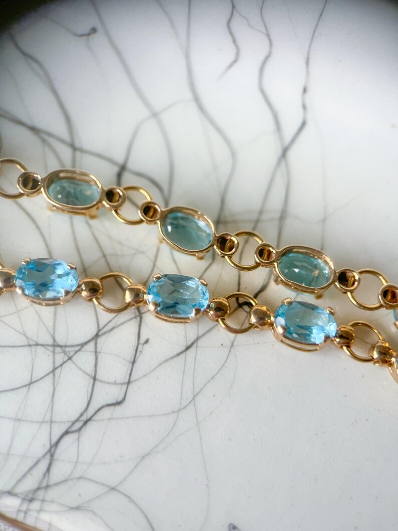 May include: Close-up of two gold-colored bracelets with oval, light blue gemstones. The gemstones are set in a gold-colored metal with small, circular accents. The bracelets are displayed on a white surface with a gray, abstract pattern.