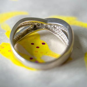 May include: A silver-toned ring with a crisscross design, featuring a band of small, clear stones. The ring is set against a backdrop of yellow and white abstract art with purple accents, creating a contrast.