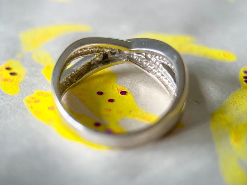 May include: A silver-toned ring with a crisscross design, featuring a band of small, clear stones. The ring is set against a backdrop of yellow and white abstract art with purple accents, creating a contrast.