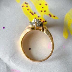 May include: A gold ring with a cluster of small, square-cut diamonds set in a square shape. The ring band is a smooth, rounded design. The background is a white surface with yellow and purple paint splatters.