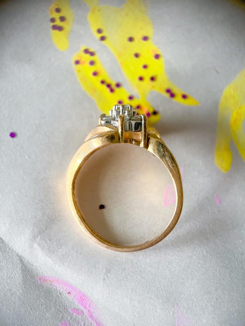 May include: A gold ring with a cluster of small, square-cut diamonds set in a square shape. The ring band is a smooth, rounded design. The background is a white surface with yellow and purple paint splatters.