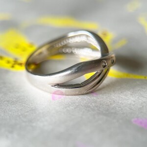 May include: A silver ring with a unique, intertwined design. The ring features a split band that crosses over itself, with small, clear stones set into the top band. The ring is displayed on a textured surface with yellow and purple accents.