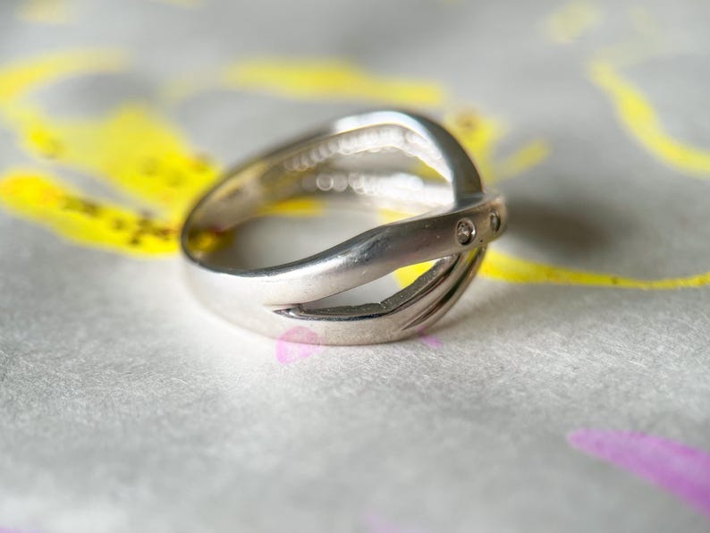 May include: A silver ring with a unique, intertwined design. The ring features a split band that crosses over itself, with small, clear stones set into the top band. The ring is displayed on a textured surface with yellow and purple accents.
