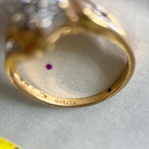 May include: Close-up of a gold ring with a cluster of small, clear stones and a small, round, red stone. The ring band is stamped with "410k". The ring is set against a white background.