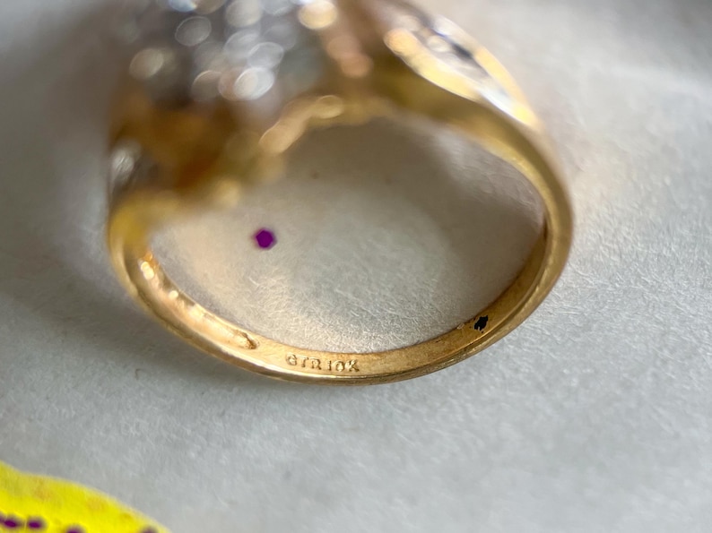 May include: Close-up of a gold ring with a cluster of small, clear stones and a small, round, red stone. The ring band is stamped with "410k". The ring is set against a white background.
