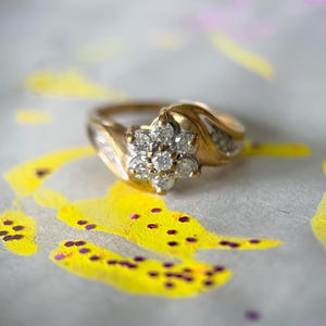 May include: A gold ring with a cluster of diamonds in a floral design. The ring is set against a background with yellow and purple paint splatters. The ring's band has a curved design.