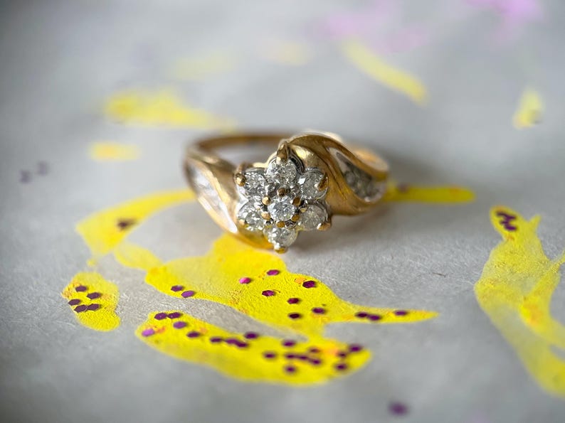 May include: A gold ring with a cluster of diamonds in a floral design. The ring is set against a background with yellow and purple paint splatters. The ring's band has a curved design.