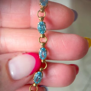 May include: A delicate gold bracelet featuring oval-cut, light blue gemstones. The gemstones are linked together with small gold rings and accents. The bracelet is held up against a blurred background.