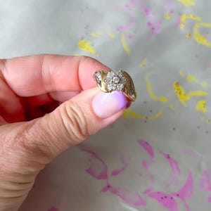 May include: A gold ring with a cluster of small, round diamonds forming a flower shape. The ring has a curved band and additional small diamonds on the sides. The ring is held against a background with pink and yellow paint splatters.