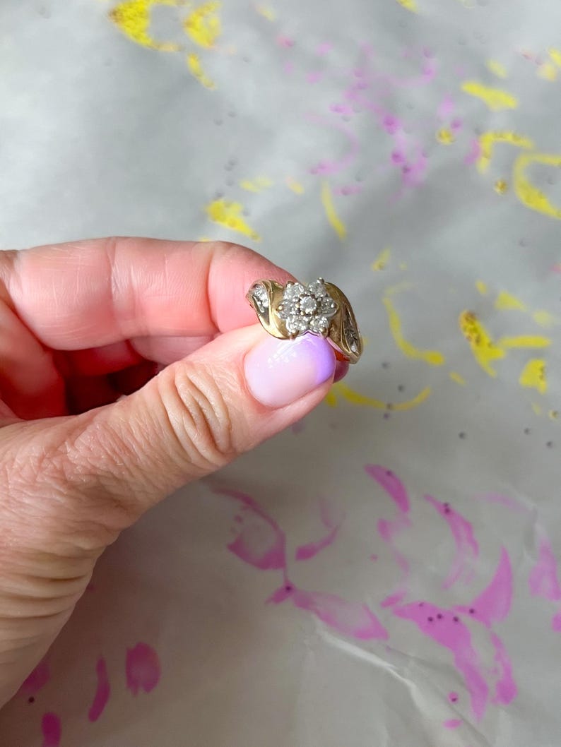 May include: A gold ring with a cluster of small, round diamonds forming a flower shape. The ring has a curved band and additional small diamonds on the sides. The ring is held against a background with pink and yellow paint splatters.