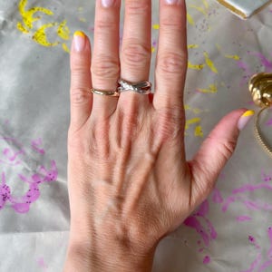 May include: A hand with a gold band ring, a silver criss-cross ring, and a gold bracelet with diamond accents. The nails are painted with a yellow French tip design.