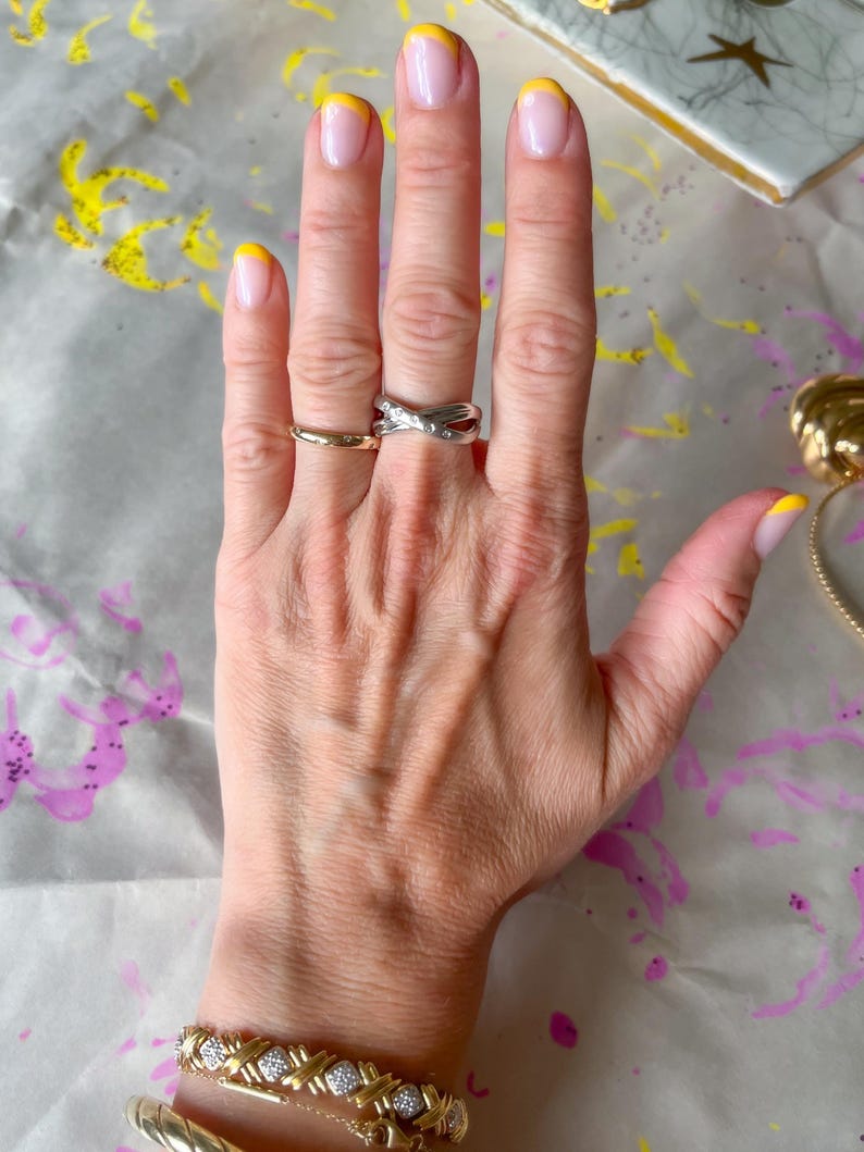 May include: A hand with a gold band ring, a silver criss-cross ring, and a gold bracelet with diamond accents. The nails are painted with a yellow French tip design.