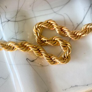 May include: Close-up of a gold-colored rope chain necklace. The chain is thick and has a twisted design. The necklace is resting on a white surface with gray markings.