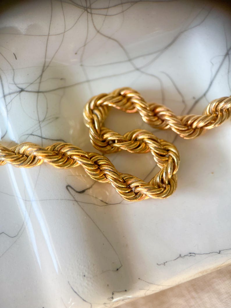 May include: Close-up of a gold-colored rope chain necklace. The chain is thick and has a twisted design. The necklace is resting on a white surface with gray markings.