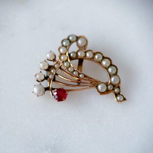 May include: A vintage gold-toned brooch featuring a cluster of small pearls and a single, vibrant red gemstone. The brooch has an ornate, flowing design, with the pearls arranged in a curved pattern. The overall aesthetic is elegant and antique.