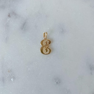 May include: A gold-colored pendant in the shape of the letter "G". The pendant is made of a shiny, reflective metal and is suspended from a small loop at the top. The background is a white marble surface.