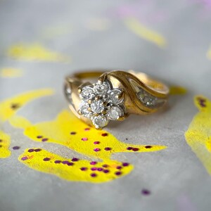 May include: A gold ring with a floral cluster of diamonds at the center. The band features a wave design with smaller diamonds. The ring is set against a background with yellow and purple paint.